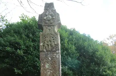 Weathered stone cross with intricate carvings, surrounded by lush greenery under a cloudy sky. Explore historical landmarks near Ardmore Country House.