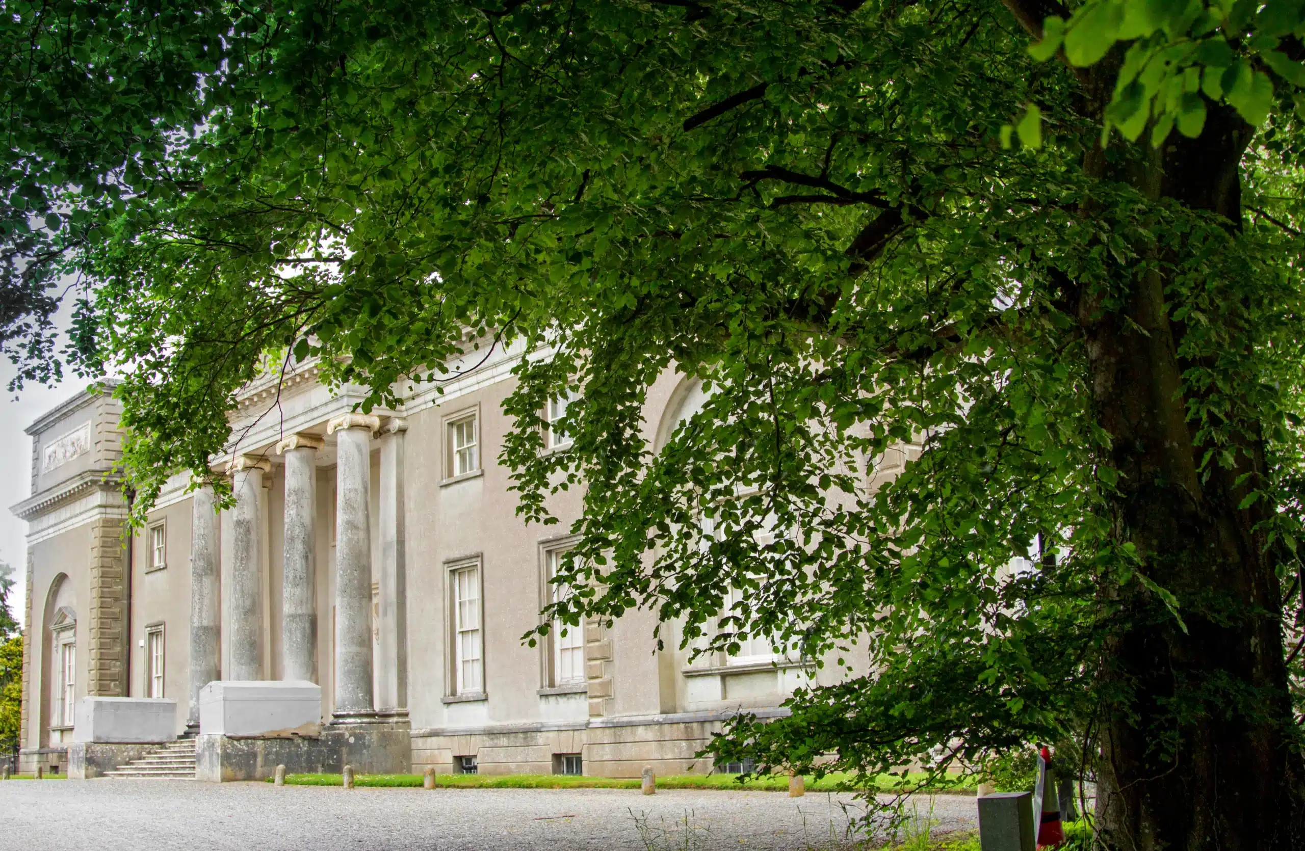 Stately building with columns, partly obscured by green tree branches, on a gravel path
