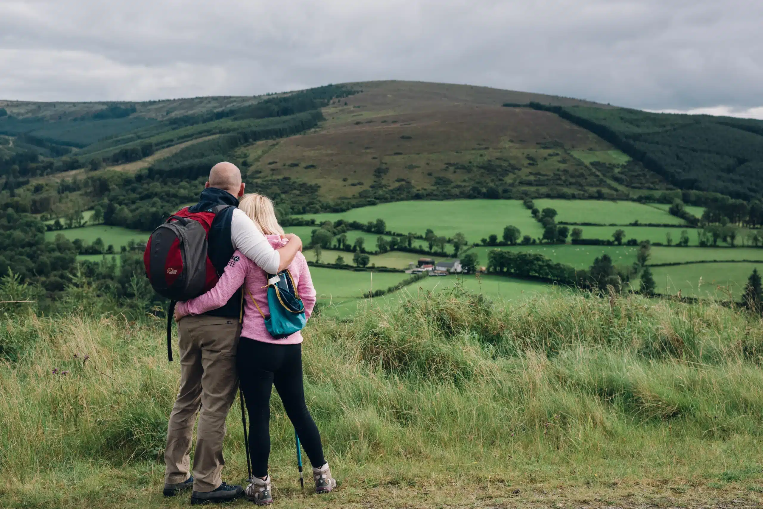 A couple embraces while overlooking a lush green valley and rolling hills under a cloudy sky during a hiking adventure at Ardmore Country House