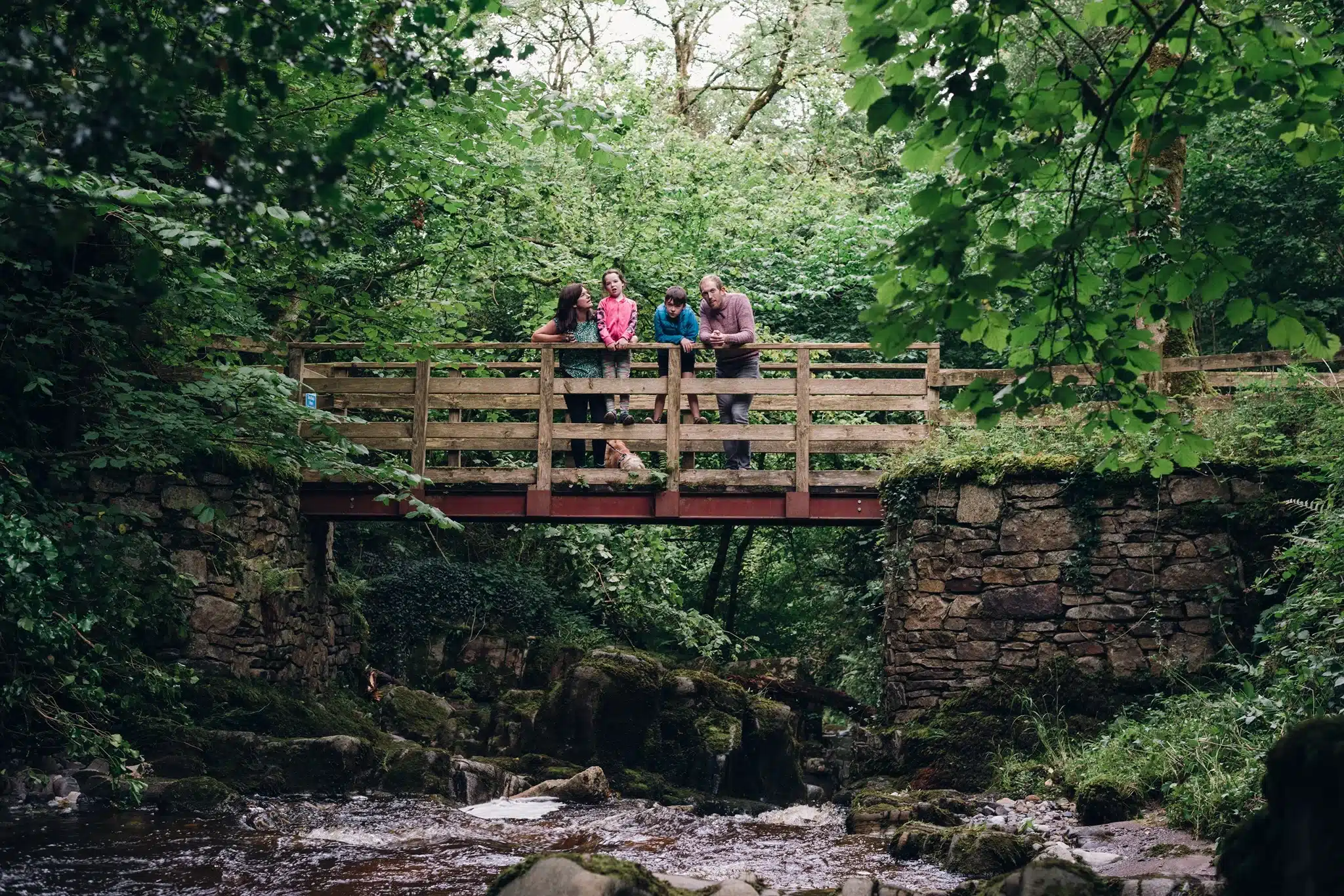A family enjoys a moment on a wooden bridge, embraced by nature with lush greenery and a peaceful river at Ardmore Country House