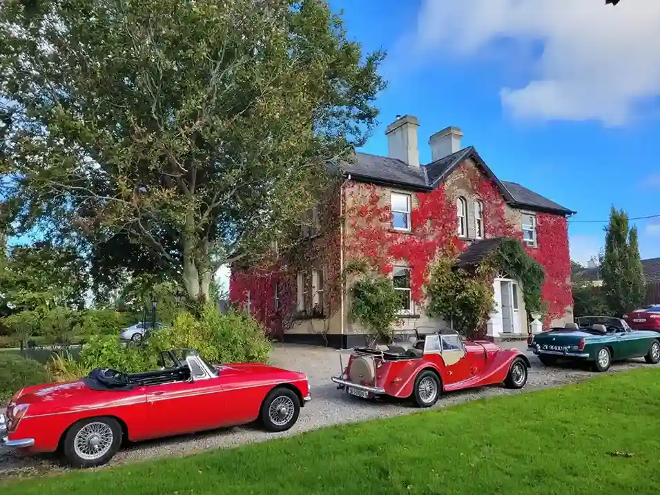 A collection of classic cars parked outside a historic home, surrounded by vibrant foliage